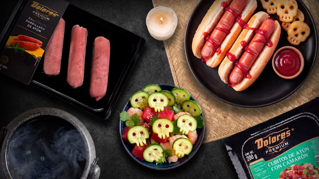 Mesa de comida temática de Halloween con hot dogs decorados como dedos sangrientos, ensalada con pepinos y papas fritas en forma de calaveras, un paquete de Salchicha Parrillera de Atún Dolores Premium y un paquete de Cubitos de Atún con Camarón Dolores Premium.